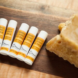 K&ouml;nnte beinhalten: F&uuml;nf wei&szlig;e Tuben mit Rosenhip- und Honig-Lippenbalsam mit einem gelben Etikett, auf dem "Rosehip & Honey Lip Balm" und "with organic rosehip seed oil" steht. Die Tuben befinden sich auf einer Holzoberfl&auml;che neben einem gro&szlig;en St&uuml;ck Bienenwachs.