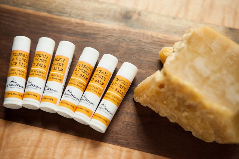 K&ouml;nnte beinhalten: F&uuml;nf wei&szlig;e Tuben mit Rosenhip- und Honig-Lippenbalsam mit einem gelben Etikett, auf dem "Rosehip & Honey Lip Balm" und "with organic rosehip seed oil" steht. Die Tuben befinden sich auf einer Holzoberfl&auml;che neben einem gro&szlig;en St&uuml;ck Bienenwachs.