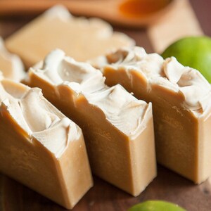 May include: Four bars of handmade soap on a wooden surface. The soap is a light brown color with white swirls. There are two green limes in the background.