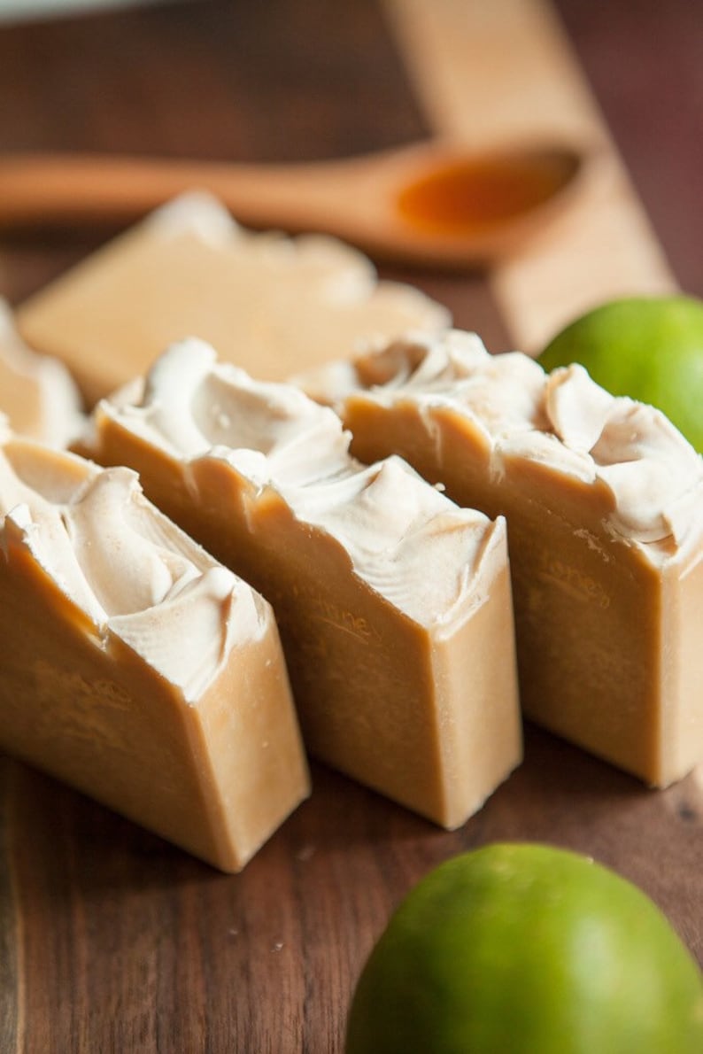 May include: Four bars of handmade soap on a wooden surface. The soap is a light brown color with white swirls. There are two green limes in the background.