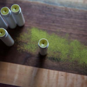 May include: Five white tubes of lip balm with yellow tops on a wooden surface. The lip balm tubes are arranged in a semi-circle with one tube in the center.