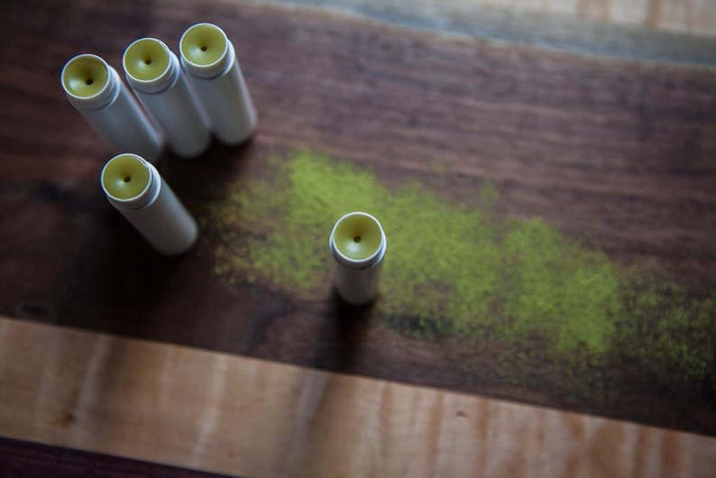 May include: Five white tubes of lip balm with yellow tops on a wooden surface. The lip balm tubes are arranged in a semi-circle with one tube in the center.