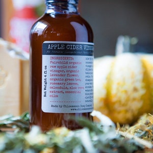 May include: A brown glass bottle with a label that reads "Apple Cider Vinegar" and lists ingredients including Fairchild organic raw apple cider vinegar, organic lavender flower, organic green tea, rosemary leaves, calendula, aloe vera extract, and essential oils. The bottle is 4 fluid ounces and is made by Chloemason Soap Company.