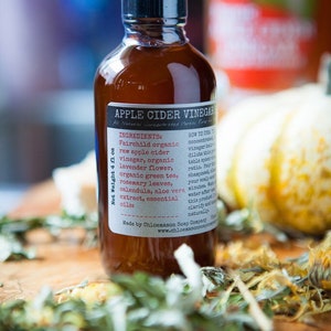 May include: A brown glass bottle with a black cap, labeled "Apple Cider Vinegar" with a list of ingredients and instructions for use. The bottle is sitting on a wooden surface with dried herbs and flowers.