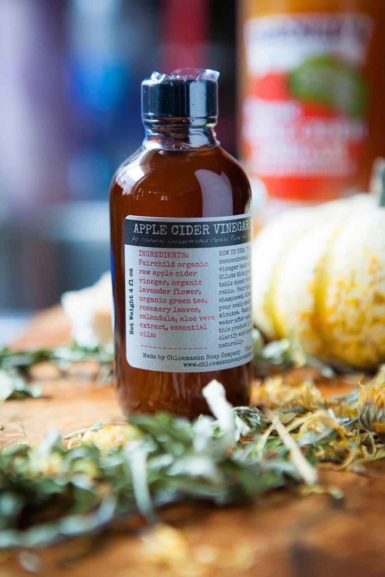 May include: A brown glass bottle with a black cap, labeled "Apple Cider Vinegar" with a list of ingredients and instructions for use. The bottle is sitting on a wooden surface with dried herbs and flowers.