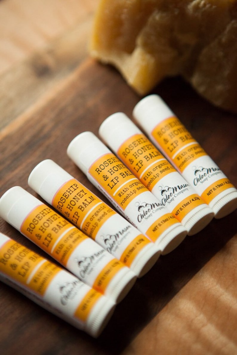 K&ouml;nnte beinhalten: Sechs wei&szlig;e Tuben mit Rosenhip- und Honig-Lippenbalsam mit gelben Etiketten. Die Etiketten tragen den Text "Rosehip & Honey Lip Balm" und "Chloe Mae Soap Company". Jede Tube ist mit einem Nettogewicht von 0,15 oz / 4,2 g gekennzeichnet.
