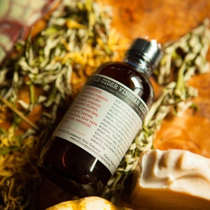 May include: A dark brown glass bottle with a white label that reads "Apple Cider Vinegar Hair Rinse" and lists ingredients including apple cider vinegar, green tea, aloe vera, and tea tree essential oil. The bottle is sitting on a wooden surface with dried flowers and a small pumpkin.