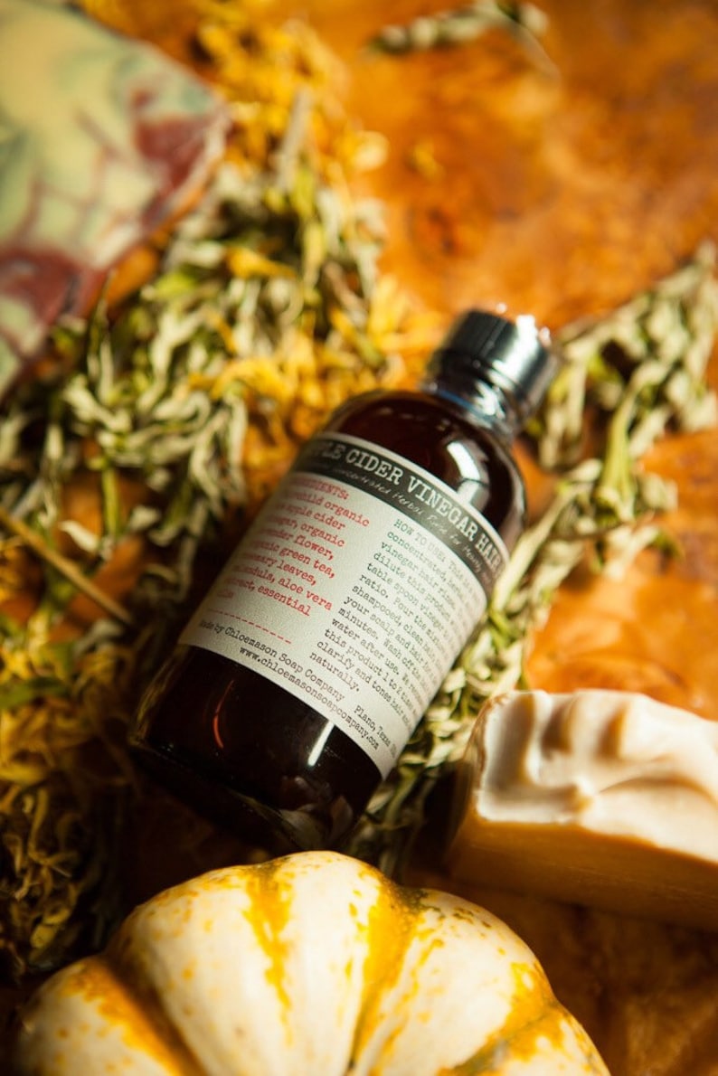 May include: A dark brown glass bottle with a white label that reads "Apple Cider Vinegar Hair Rinse" and lists ingredients including apple cider vinegar, green tea, aloe vera, and tea tree essential oil. The bottle is sitting on a wooden surface with dried flowers and a small pumpkin.