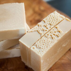 Pu&ograve; includere: Una pila di tre saponette beige lisce e una singola saponetta beige con un delicato motivo a pizzo impresso sulla parte superiore.