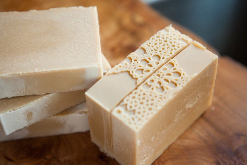 Pu&ograve; includere: Una pila di tre saponette beige lisce e una singola saponetta beige con un delicato motivo a pizzo impresso sulla parte superiore.