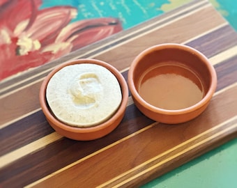 May include: Two small, round, terracotta-colored ceramic bowls. One bowl is filled with a textured, off-white substance, while the other bowl is empty, revealing a smooth, glossy, light brown interior surface.