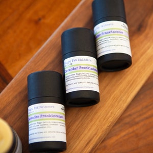 May include: Three black tubes of lavender frankincense balm with white labels. The labels have the text "Magnu Fat Skincare" and "Lavender Frankincense" on them. The tubes are on a wooden surface.