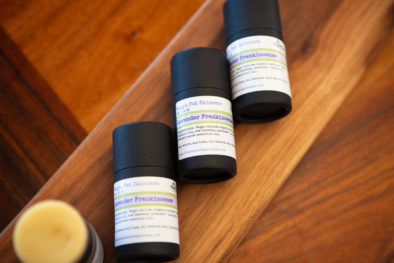 May include: Three black tubes of lavender frankincense balm with white labels. The labels have the text "Magnu Fat Skincare" and "Lavender Frankincense" on them. The tubes are on a wooden surface.