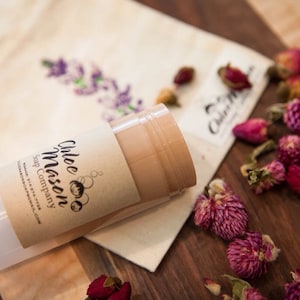May include: A white tube of natural deodorant with a brown label that says "Chloe Mason Soap Company" and features a cartoon illustration of a woman with a beehive hairstyle. The tube is sitting on a white cloth bag with dried flowers scattered around it.