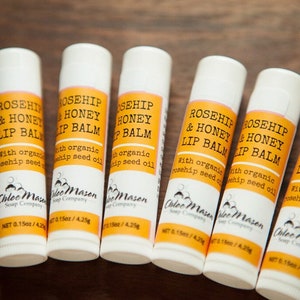 K&ouml;nnte beinhalten: Sechs wei&szlig;e Tuben mit Rosehip- und Honig-Lippenbalsam mit gelben Etiketten. Die Etiketten lauten "Rosehip & Honey Lip Balm" mit dem Text "With organic rosehip seed oil" darunter. Die Tuben sind in einer Reihe auf einer Holzoberfl&auml;che angeordnet.