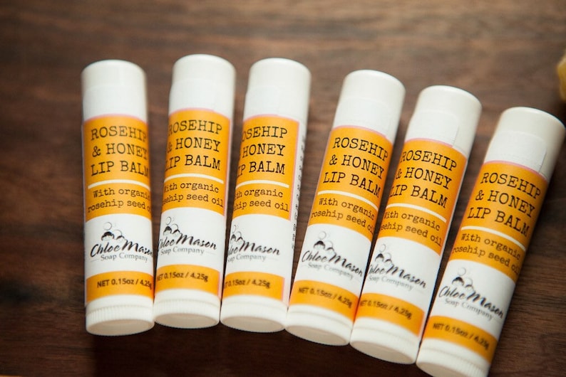 K&ouml;nnte beinhalten: Sechs wei&szlig;e Tuben mit Rosehip- und Honig-Lippenbalsam mit gelben Etiketten. Die Etiketten lauten "Rosehip & Honey Lip Balm" mit dem Text "With organic rosehip seed oil" darunter. Die Tuben sind in einer Reihe auf einer Holzoberfl&auml;che angeordnet.
