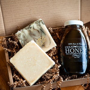 May include: A gift box filled with brown packing paper and two bars of soap, one white and one green, and a glass jar of Texas Orange Honey. The honey jar has a white label with the text "100% Raw & Natural Texas Orange Honey Raw and Unfiltered Texas Honey 16oz / 454g"