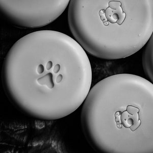 May include: Five round, white, ceramic soap dishes with different animal paw prints and dog silhouettes. The soap dishes are on a dark wood surface.