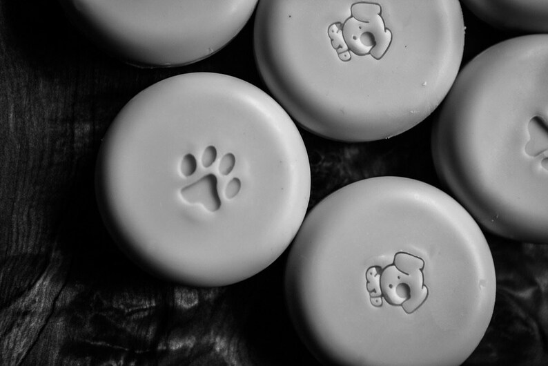May include: Five round, white, ceramic soap dishes with different animal paw prints and dog silhouettes. The soap dishes are on a dark wood surface.