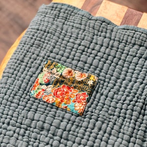 May include: A gray quilted kitchen towel with a floral fabric patch sewn on. The towel has a textured, waffle-like pattern.