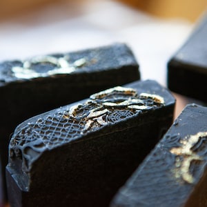 May include: Close-up of several rectangular black soaps with gold and silver lace-like designs on the top. The soaps are arranged in a slightly scattered pattern, with a shallow depth of field.