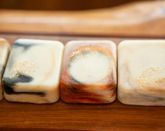 May include: A row of rectangular soap bars with rounded edges. Each bar features a unique marbled pattern in shades of cream, black, and orange. Some bars are embossed with a delicate floral stamp, while others show smooth surfaces.