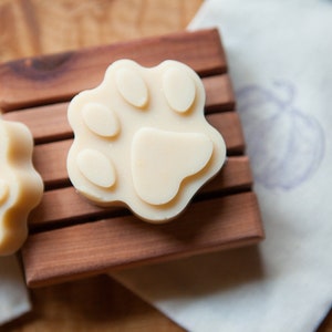 May include: Three white paw-shaped soaps on a wooden soap dish. The soaps are made with natural ingredients and are ideal for sensitive skin.