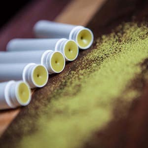 May include: Five white tubes of lip balm with yellow balm inside, arranged in a row on a wooden surface with green powder scattered around them.