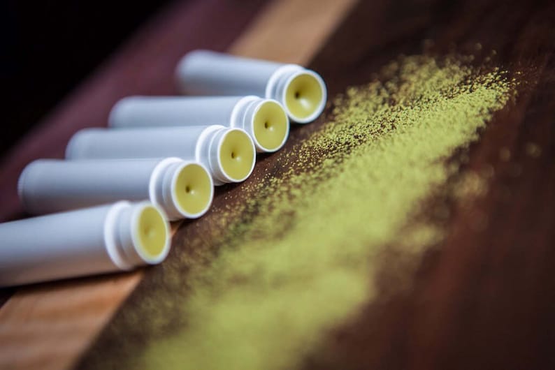 May include: Five white tubes of lip balm with yellow balm inside, arranged in a row on a wooden surface with green powder scattered around them.