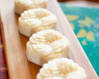 May include: Four round, off-white soap bars arranged in a single line. Each soap features a detailed, raised floral pattern on its top surface, consisting of multiple layered, curved petals. The soaps have a smooth, matte texture and a uniform color.