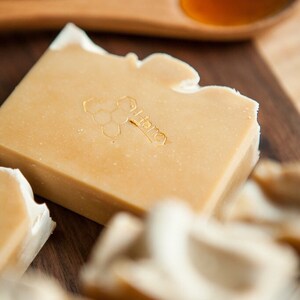 May include: A close-up of a bar of honey soap with a honeycomb design and the word "Honey" embossed on the top. The soap is on a wooden surface with a wooden spoon filled with honey.