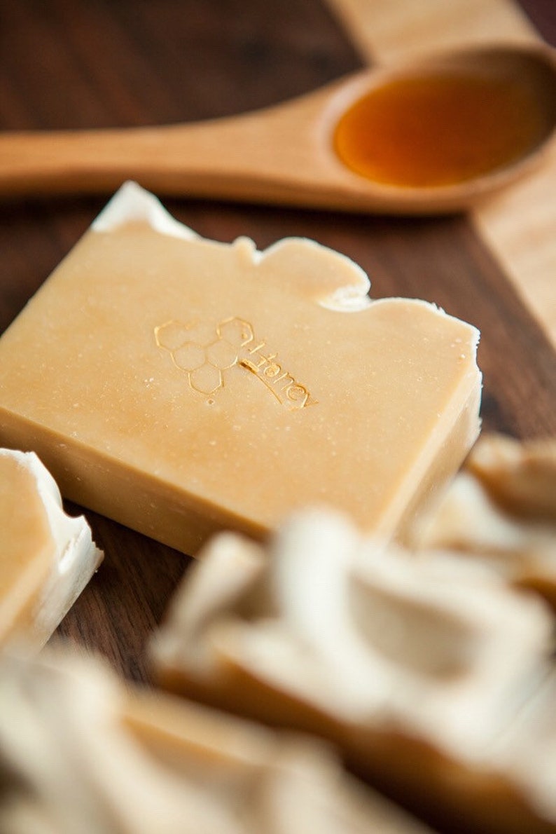 May include: A close-up of a bar of honey soap with a honeycomb design and the word "Honey" embossed on the top. The soap is on a wooden surface with a wooden spoon filled with honey.