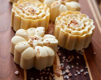 May include: Several flower-shaped, light-colored soaps with a textured surface are arranged on a wooden cutting board. Some soaps have a sprinkle of small, light-brown granules on top. The wooden board has dark and light brown stripes.