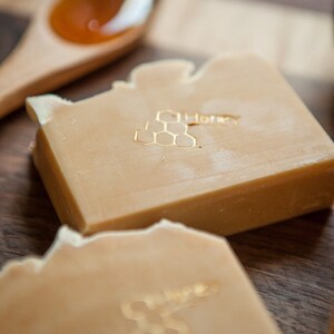May include: Two bars of honey soap with a honeycomb design and the word "Honey" embossed on the top. The soap is a light beige color and is sitting on a wooden surface.