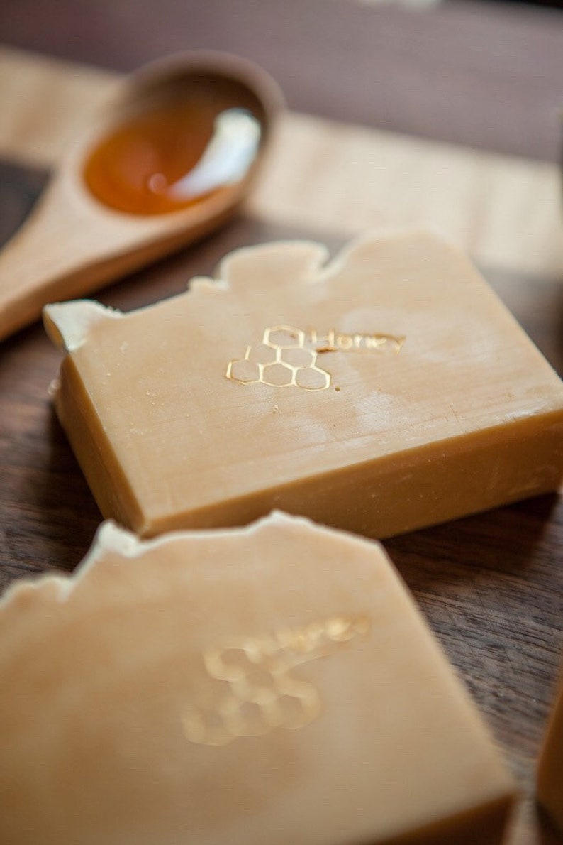 May include: Two bars of honey soap with a honeycomb design and the word "Honey" embossed on the top. The soap is a light beige color and is sitting on a wooden surface.