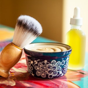 May include: A blue and white ceramic bowl filled with shaving cream, a wooden shaving brush with white bristles, and a clear glass bottle containing yellow liquid.