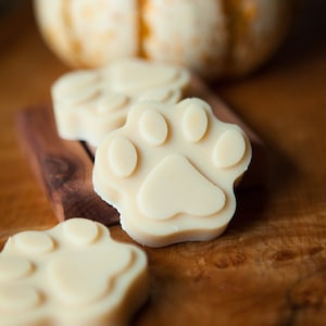 May include: Three white paw-shaped soaps on a wooden surface. The soaps are made in the shape of a paw print and are a light cream colour.