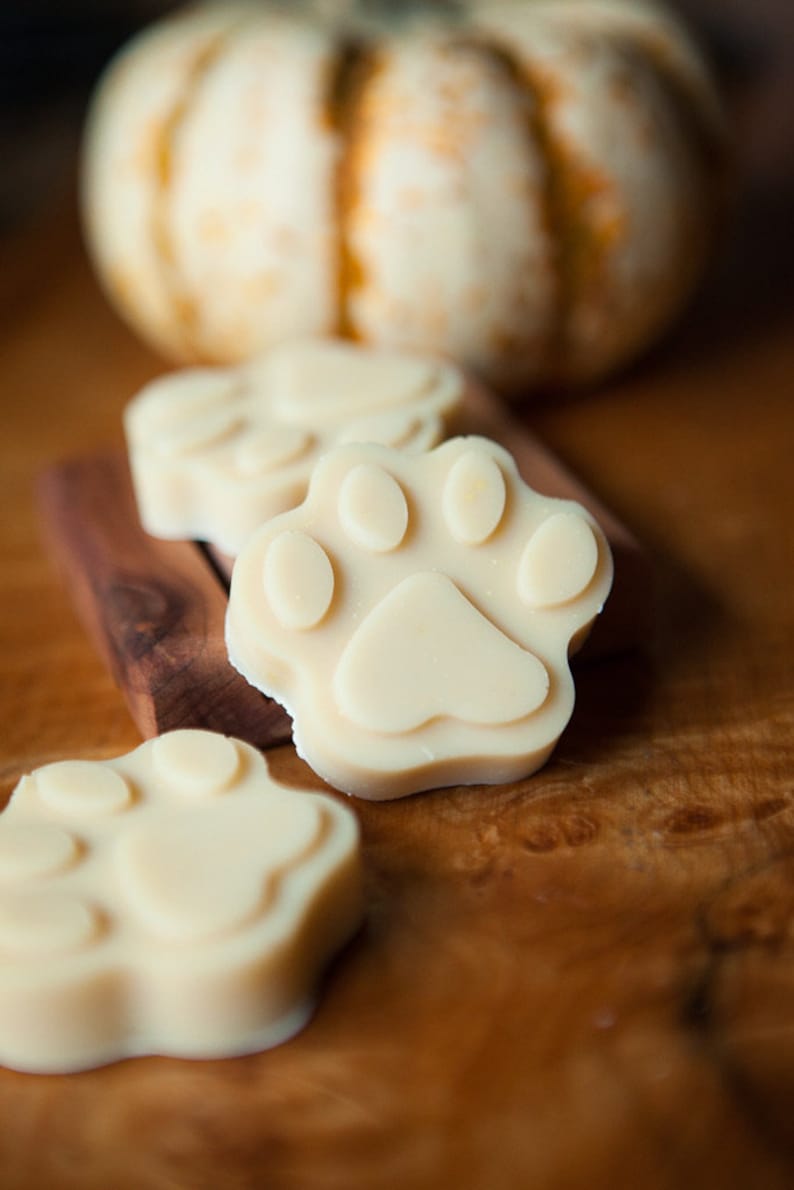 May include: Three white paw-shaped soaps on a wooden surface. The soaps are made in the shape of a paw print and are a light cream colour.