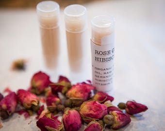 May include: Three clear tubes of lip balm with a white label on the front tube. The label reads "ROSE GERANIUM &HIBISCUS" and lists the ingredients: "ORGANIC OIL, RAW GERANIUM, HIBISCUS, VITAMIN E". Dried rose buds are scattered around the tubes.