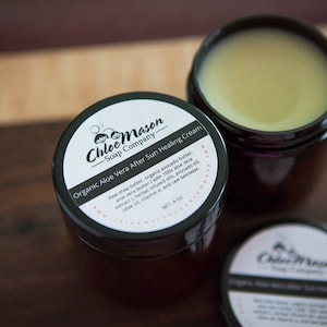 May include: Two black jars of organic aloe vera after sun healing cream from Chloe Mason Soap Company. The jars have black lids and white labels with the company name and product description. The jars are sitting on a wooden surface.