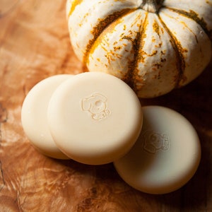 May include: Three round, white, unscented soap bars with a small dog logo on each bar. The soap bars are sitting on a wooden surface next to a small, white and orange pumpkin.