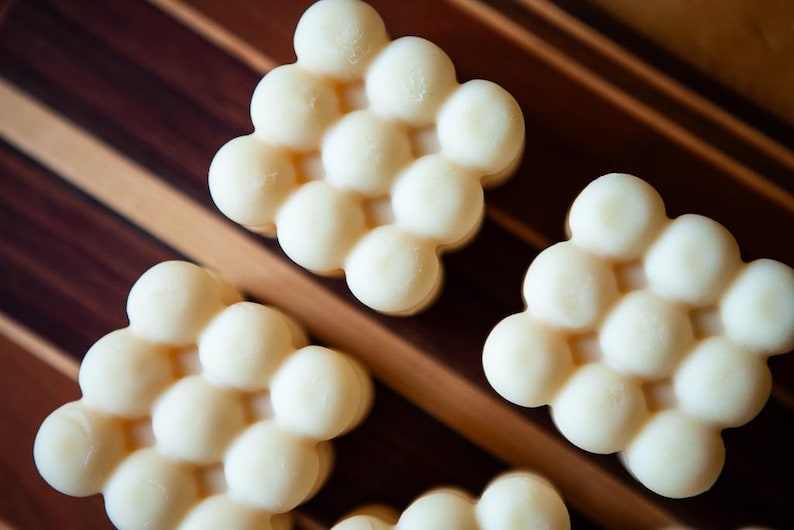 May include: Close-up of several off-white, honeycomb-shaped soaps. The soaps are arranged on a wooden surface with brown and tan stripes. The soaps have a smooth, rounded texture.