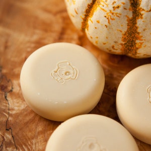 May include: Three round, white, unscented soap bars with a small dog logo on each bar. The soap bars are sitting on a wooden surface next to a small white pumpkin.