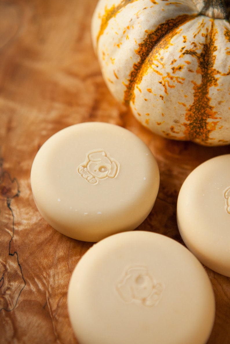 May include: Three round, white, unscented soap bars with a small dog logo on each bar. The soap bars are sitting on a wooden surface next to a small white pumpkin.