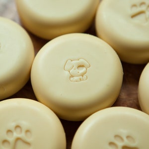 May include: Six round, pale yellow soap bars with different animal paw prints embossed on the top. The soap bars are arranged in a semi-circle on a wooden surface.