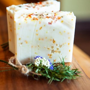May include: Two white, square bars of soap with a star-shaped design cut into the front. The soap is speckled with orange and brown flecks and topped with herbs. A bundle of greenery and flowers is tied with twine in front.