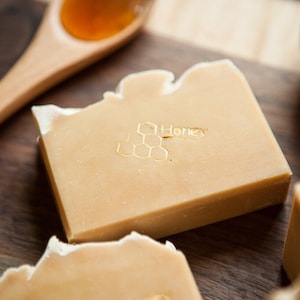 May include: Two bars of honey soap with a honeycomb design and the word "Honey" printed on them. The soap is on a wooden surface with a wooden spoon filled with honey.