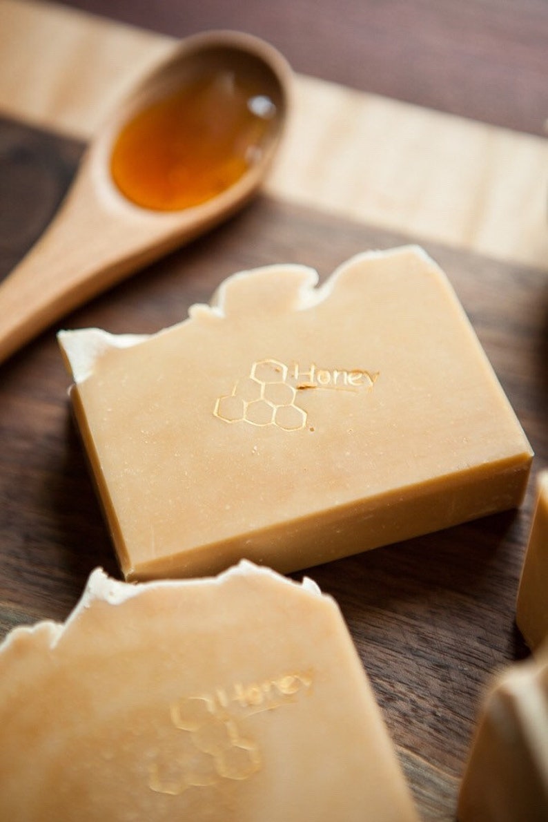 May include: Two bars of honey soap with a honeycomb design and the word "Honey" printed on them. The soap is on a wooden surface with a wooden spoon filled with honey.