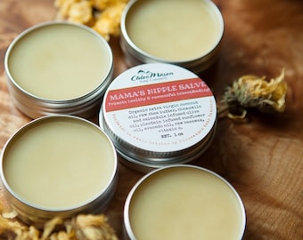 May include: Close-up of several small, round metal tins filled with a pale yellow balm. One tin has a label that reads "Mama's Nipple Salve" with a list of ingredients. Dried yellow flowers are scattered around the tins on a wooden surface.