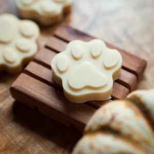 May include: Four white paw-shaped soaps on a wooden soap dish. The soaps are made in the shape of a dog's paw print.
